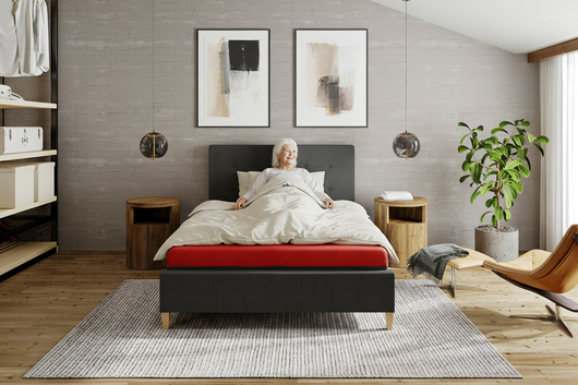 Woman resting on a red ThevoautoActiv pressure-care mattress with white bedding in a modern bedroom.