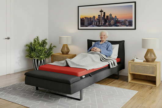 Man reclining on a red ThevoautoActiv pressure-care mattress in a raised adjustable bed with a gray blanket.