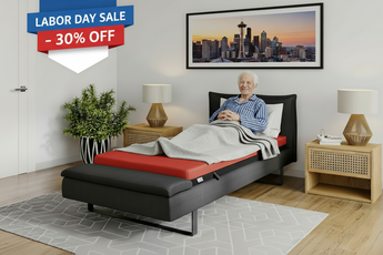 A modern adjustable bed with a black upholstered frame and red mattress, shown in a styled bedroom with an older man lying down. The scene includes a Labor Day Sale banner overlay, a cityscape print on the wall, bedside tables, lamps, and a plant.