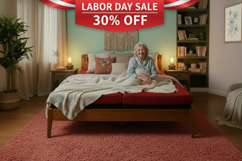 A woman sits on a wooden bed with a red mattress and a cream knit blanket in a styled bedroom. The room has a mint green wall, a macrame wall hanging, a tall bookshelf, and a large pinkish-red textured rug.