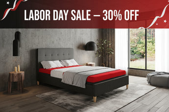 A modern bed with a dark gray upholstered frame and light wood legs, dressed in red and white bedding, set in a contemporary bedroom with a concrete wall, large window, and gray rug.
