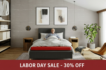A man lies in bed on a modern dark gray upholstered bed frame with a red mattress edge, in a styled bedroom with two abstract wall art prints, wooden nightstands, a plant, and a chair.